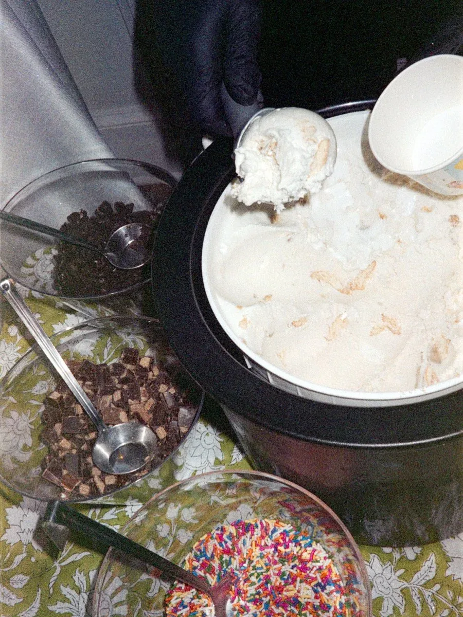 A tub of vanilla ice cream at a sundae bar, with bowls of chocolate chips, sprinkles, and other toppings