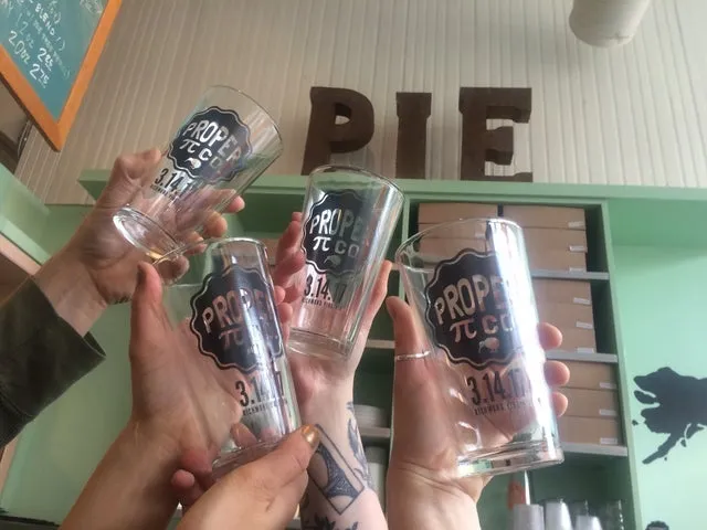 Hands raising Proper Pie pint glasses in front of the shop’s mint-green shelves and big PIE letters