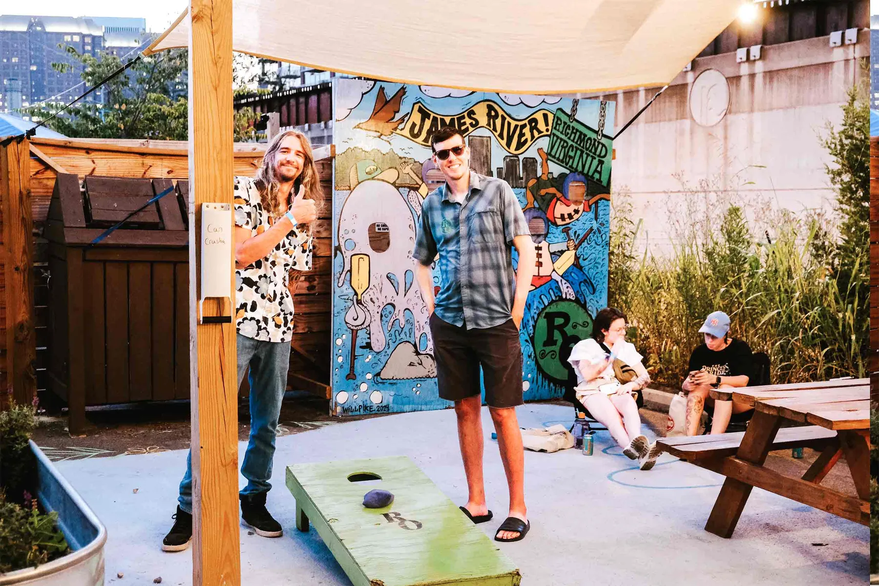 Friends relaxing on the Riverside shop patio beside a river mural and a cornhole board
