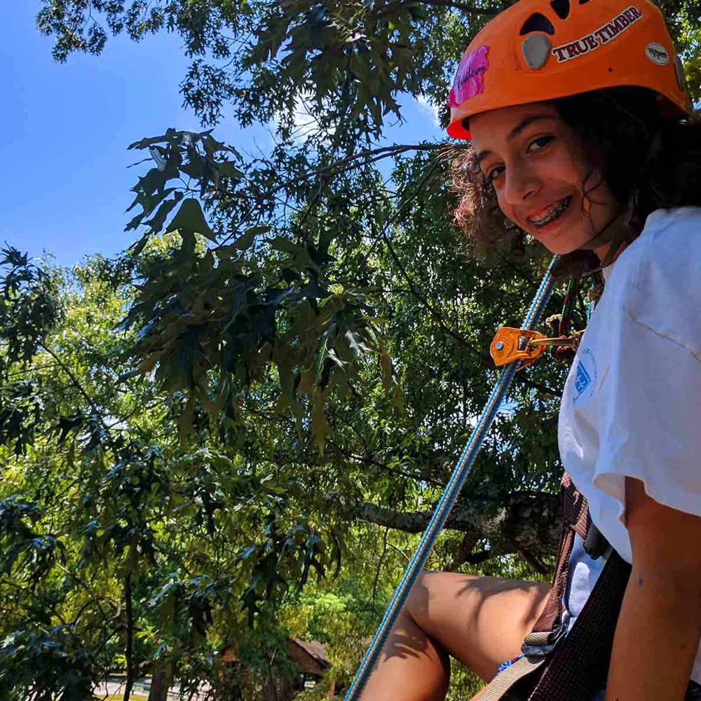 A smiling camper in a helmet and harness climbing a tree