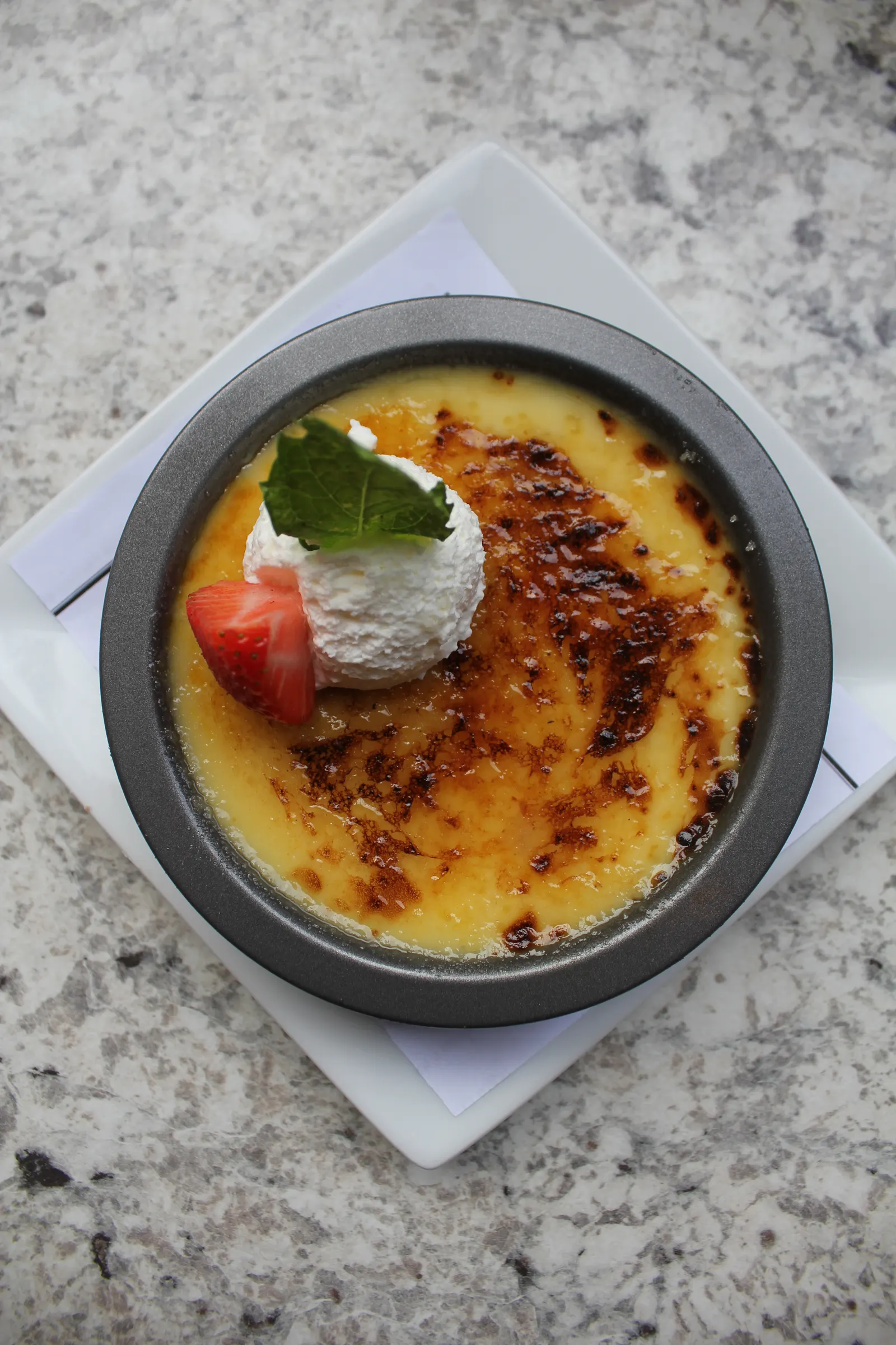 Crème brûlée in a dark ramekin with a torched top, whipped cream, a strawberry, and mint