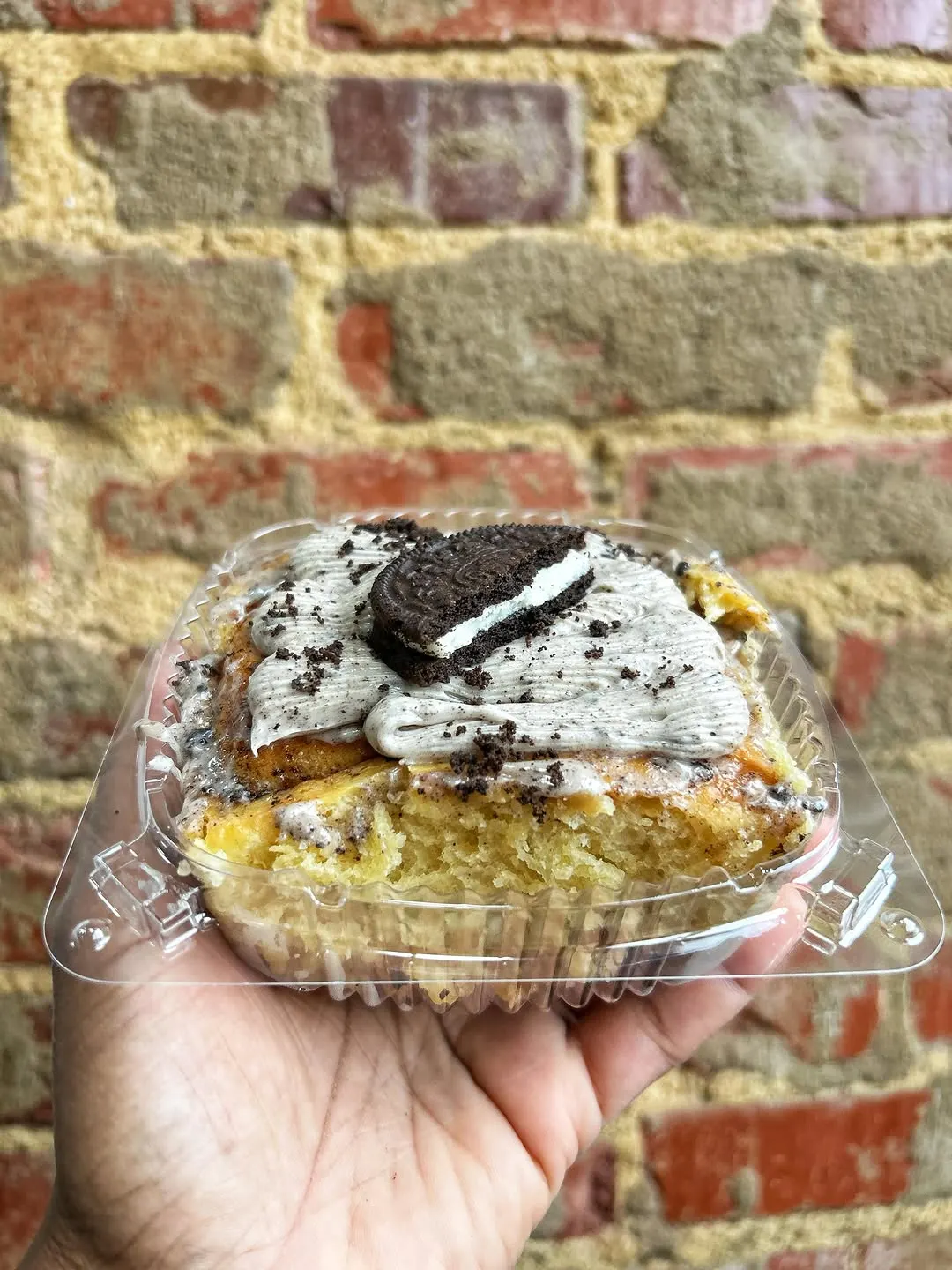 A hand holding a clear to-go box of a cookies-and-cream dessert against a brick wall