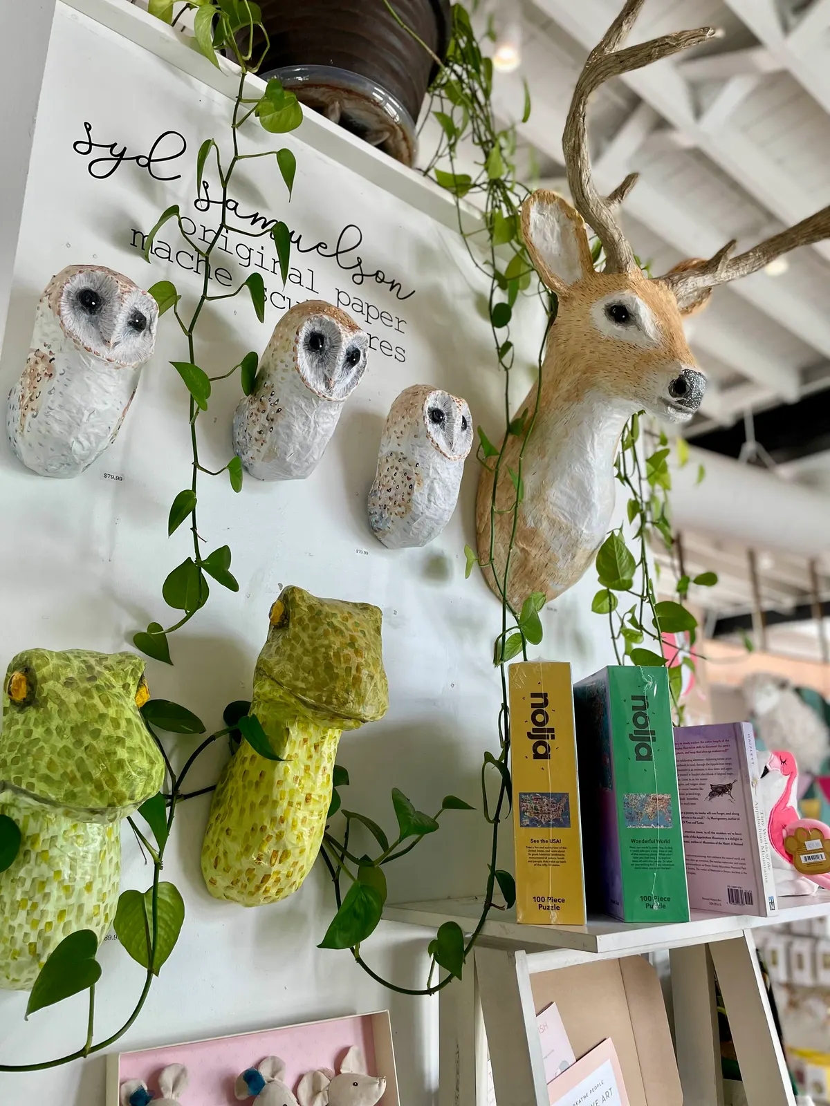 Papier-mâché owls, frogs, and a deer head on a white gift shop wall with trailing vines
